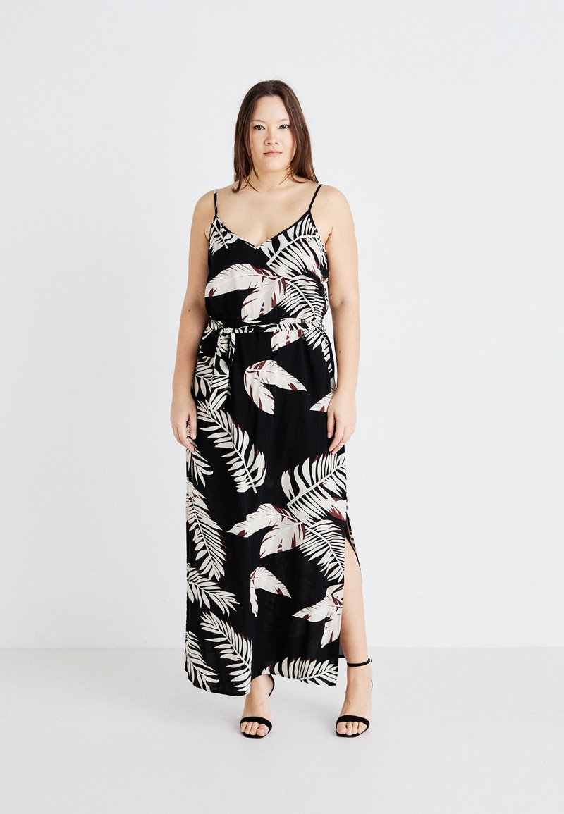 Robe maxi noire avec un motif de feuilles blanc et beige, décolleté en v, bretelles fines, taille marquée et fente latérale. Modèle portant des sandales noires à brides.