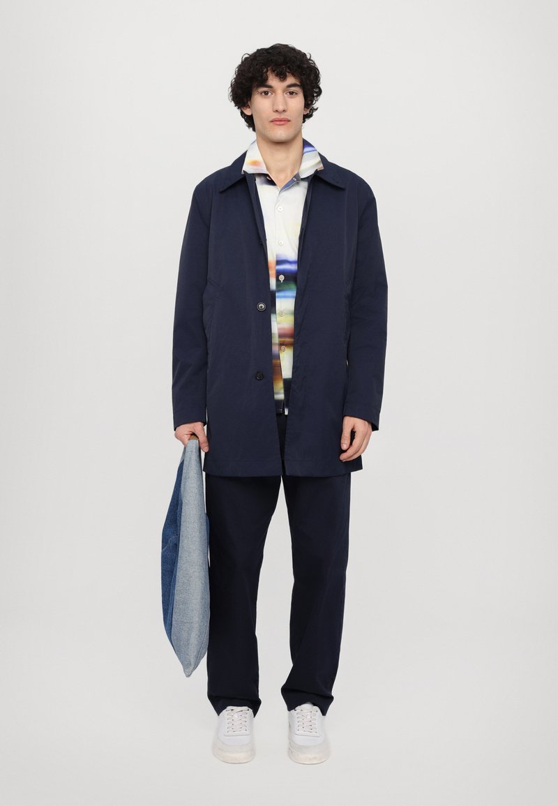 Marineblauer Trenchcoat mit geradem Schnitt, über einem mehrfarbigen gestreiften Hemd getragen, kombiniert mit dunklen Hosen und weißen Sneakern, während eine Denim-Tasche gehalten wird.