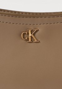 Brunlædertaske med et gyldent "cK" logo, glat tekstur og fine syninger langs kanterne. Minimalistisk design med et moderne udseende.
