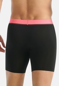 Man in zwarte zwembroek met een roze tailleband, gezien vanuit de achterkant tegen een witte achtergrond.