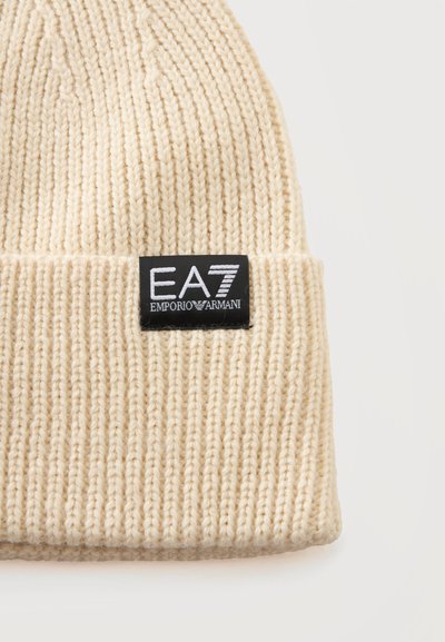 Bonnet tricoté de couleur crème avec un revers, présentant une étiquette noire avec la marque blanche EA7 Emporio Armani brodée.