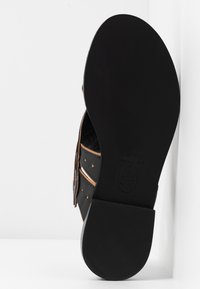 Sandale en cuir noir avec un design à découpes, des coutures contrastantes en marron et des accents de clous ronds. Semelle plate noire avec une texture lisse.