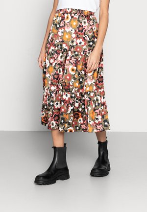 Femme portant une jupe midi fleurie colorée associée à des bottines noires, debout contre un mur clair et un sol uni.