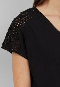 Haut en coton noir avec un décolleté en V et des manches courtes. Les manches présentent des détails en dentelle noire délicate, offrant un contraste texturé.