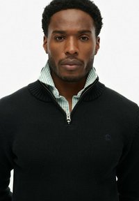 Czarny sweter z dzianiny z wysokim kołnierzem i zamkiem, założony na jasnoszaro-biały pasek. Gładka tekstura.