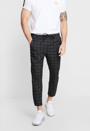 Homme portant un t-shirt blanc, un pantalon à cordon noir à carreaux et des baskets blanches, debout les mains dans les poches sur un fond uni.