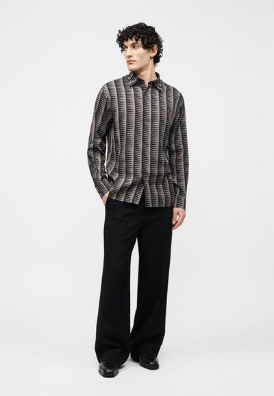 Νεαρός με σγουρά μαλλιά, φορώντας πουκάμισο με γεωμετρικό σχέδιο και μαύρο παντελόνι φαρδύ, στέκεται με τα χέρια στις τσέπες.