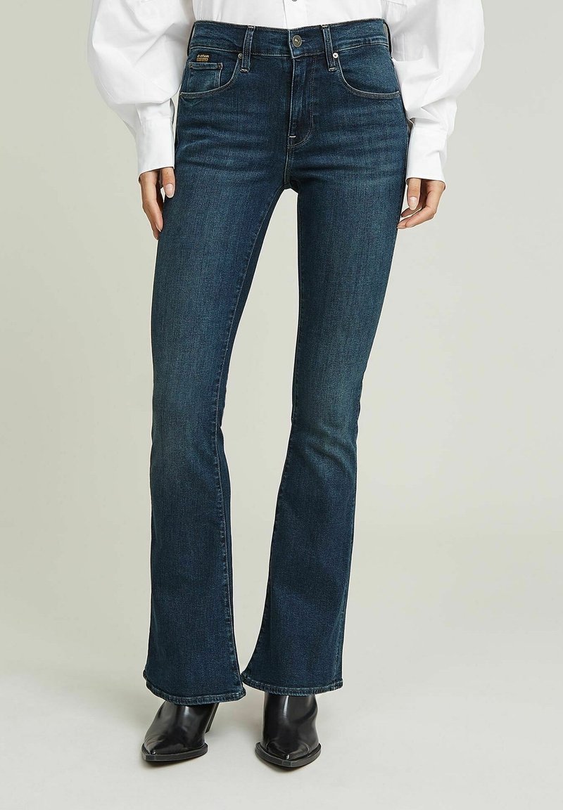 Jean évasé bleu foncé porté avec des bottes pointues noires et une chemise blanche à manches longues, montré de la taille aux pieds.