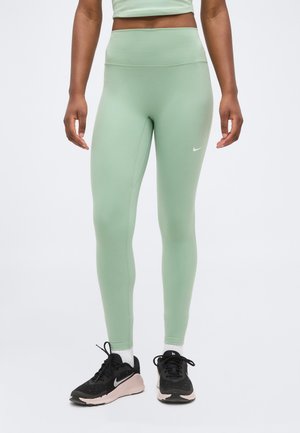 Person iført lysegrønne leggings med høj talje og sorte Nike-sneakers med hvide sokker, stående mod en hvid baggrund.