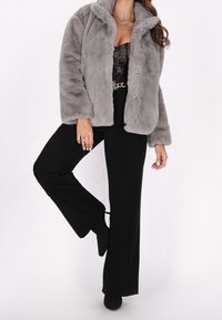 Giacca in pelliccia sintetica grigia, taglio ampio, con collo. Indossata sopra un top in pizzo nero e pantaloni neri a vita alta, leggermente svasati.