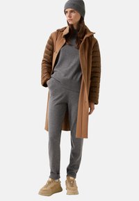 Kvinde iført grå strikhue og matchende sweater og bukser, camelbrun jakke med polstrede ærmer og beige sneakers, står med den ene hånd i lommen.