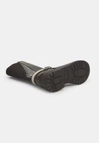Bota negra de ante hasta la rodilla con suela texturizada. Cuenta con una correa adornada con strass y un diseño de punta afilada.