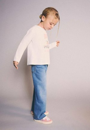 Cămașă albă cu mâneci lungi cu grafică colorată, blugi din denim albastru și pantofi slip-on roz pastel. Coafură simplă cu o împletitură.