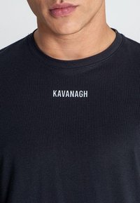 Crni rebrasti majica s okruglim izrezom, s nazivom branda "KAVANAGH" u bijeloj boji, centriranom na prsima. Mekana tkanina.