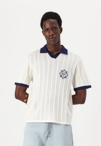 Polo de punto blanco con cuello y puños en azul marino. Presenta un parche con logo y un patrón de rayas verticales. Diseño informal de manga corta.