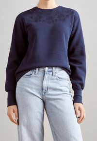 Felpa navy con colletto rotondo, decorata con motivi floreali ricamati lungo le spalle. Abbinata a jeans azzurri a vita alta.