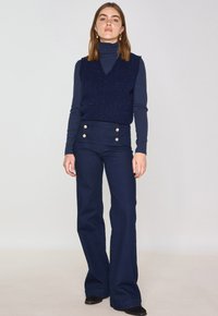 Marineblauer Weste mit strukturiertem Finish, die über einem eng anliegenden Rollkragenpullover getragen wird, kombiniert mit hoch taillierten, ausgestellten Marineblauen Hosen mit goldenen Knöpfen.