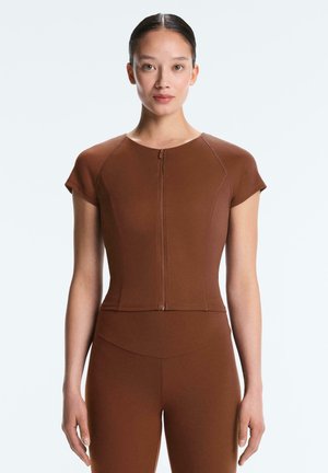 Donna con indosso un top marrone aderente a maniche corte con cerniera e leggings coordinati a vita alta, in piedi davanti a uno sfondo bianco semplice.