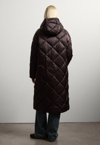 Giacca piumino marrone trapuntata con cappuccio, caratterizzata da un motivo a diamante. La giacca arriva fino a metà polpaccio, abbinata a pantaloni di denim larghi.