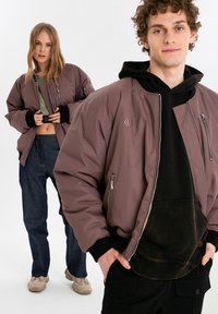 Brun bomberjacka med dragkedjefickor och ribbade muddar, bärs över en svart hoodie. Modell i förgrunden, en annan modell delvis synlig bakom.