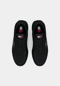 Black sneakers with a mesh upper, featuring a textured sole, tonal laces, and a striped logo tag in red, white, and blue on the tongues.
