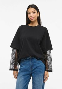 Svart oversized t-skjorte med lange ermer i blonde. Mykt bomullsmateriale, rund halslinning og avslappet passform kombinert med blå denimjeans.