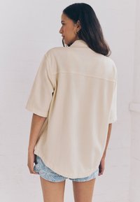 Camicia beige oversize a maniche corte con cucitura sul dietro, dal taglio rilassato e leggermente testurizzata, abbinata a pantaloncini blu con motivo a fantasia.