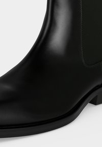 Gros plan d'une bottine en cuir noir lisse avec un panneau latéral élastique et un petit talon sur un fond uni.