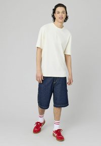 T-shirt crème à manches courtes associé à un short en denim bleu. Le modèle porte des baskets rouges et des chaussettes blanches avec des rayures rouges. Design simple et coupe décontractée.