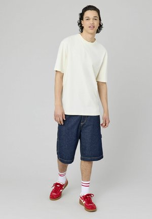T-shirt crème à manches courtes associé à un short en denim bleu. Le modèle porte des baskets rouges et des chaussettes blanches avec des rayures rouges. Design simple et coupe décontractée.