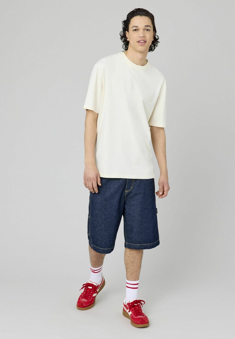 T-shirt crème à manches courtes associé à un short en denim bleu. Le modèle porte des baskets rouges et des chaussettes blanches avec des rayures rouges. Design simple et coupe décontractée.