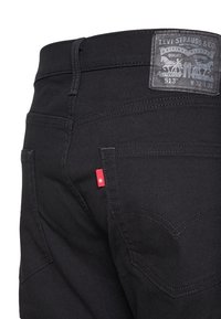 Black denim jeans with a fitted design, featuring a dark leather patch, rear pocket stitching in a curved pattern, and a small red logo tab.