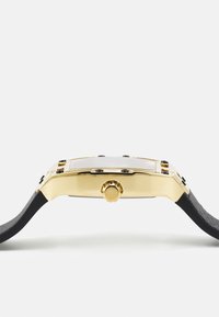 Kuldtooniline kell, millel on ristkülikuline korpus, must tekstuuritud rihm ja silmapaistev kroon, selge näo ning lihtsate joonte ja mustade märkidega.