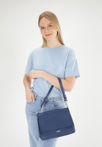 Een marineblauwe schoudertas met een gestructureerd oppervlak, een klepafsluiting en een verstelbare schouderriem, gecombineerd met een lichtblauw shirt en denim jeans.
