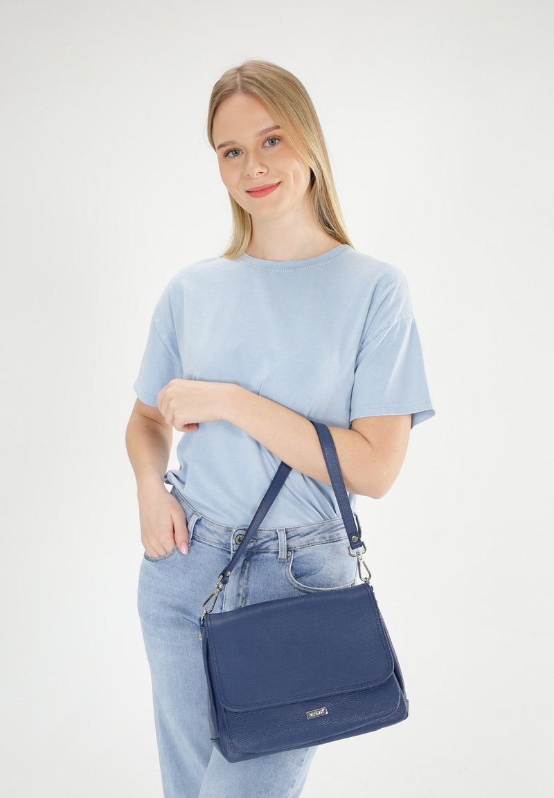 Een marineblauwe schoudertas met een gestructureerd oppervlak, een klepafsluiting en een verstelbare schouderriem, gecombineerd met een lichtblauw shirt en denim jeans.