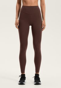 Smeđe legice s visokim strukom izrađene od rastezljivog materijala, s glatkom teksturom i pristajućim dizajnom koji seže do gležnjeva.