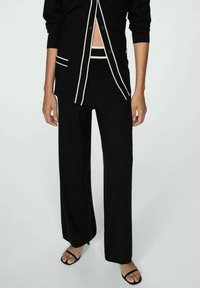 Gilet en tricot noir avec des accents blancs, associé à un pantalon noir à jambes larges et des sandales à talons noires. Le gilet se ferme par des boutons.