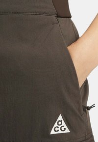 Mörkbruna tygshorts med en triangelformad vit logotyp. Funktioner inkluderar sidofickor, strukturerat material och justering med dragsko.