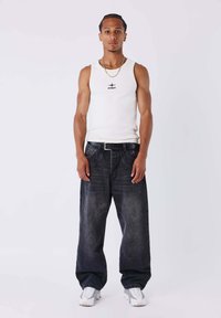 Canotta bianca senza maniche con logo nero, abbinata a jeans blu scuro ampi e scarpe sportive bianche. Design minimalista e vestibilità comoda.