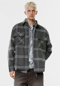 Šedá károvaná košele s gombíkmi, s dvoma náprsními vreckami, vrstvená nad károvanou košeľou. Mäkká látka s voľným strihom.