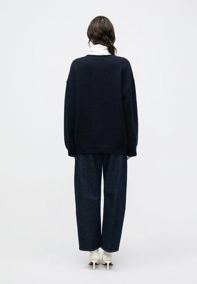 Suéter de punto extragrande color azul marino, tela texturizada, puños y dobladillo acanalados, combinado con pantalones vaqueros de pierna ancha y botines de tacón.