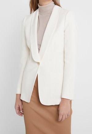 Femme portant un blazer blanc à un bouton sur un col roulé beige et une jupe couleur camel, debout devant un fond uni.