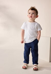 Barn iført en hvid bomulds T-shirt, marineblå linnedbukser med op rullede manchetter og hvide sandaler med justerbare stropper.