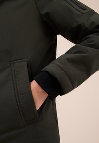 Veste matelassée vert foncé avec une poche à fermeture éclair sur l'épaule, des poignets en tricot côtelé et une poche latérale avec des détails de couture verticale.