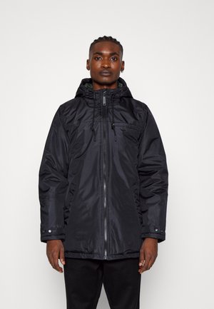 Chaqueta negra con capucha, cremallera y cordones, presenta una textura suave, puños elásticos y un ligero brillo, usada sobre pantalones negros.