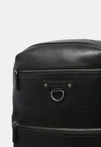 Zaino in pelle nera con finiture in argento, dotato di un compartimento principale con zip, tasca anteriore con zip e un'etichetta di marca con dettaglio ad anello metallico.