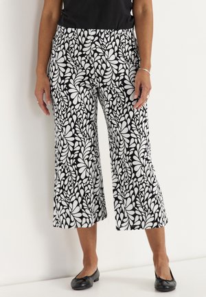 Kobieta ubrana w czarno-białe wzorzyste szerokie spodnie 3/4, czarny top, czarne baleriny i srebrną bransoletkę, stoi przy białej ścianie.