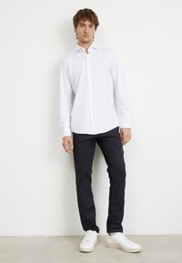 Camicia bianca a maniche lunghe con colletto, abbinata a pantaloni scuri e sneakers bianche. Design semplice con una texture liscia.
