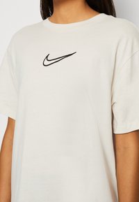 Krämfärgad bomulls-T-shirt med korta ärmar, med en svart Nike-logotyp broderad på bröstet. Mjuk textur och klassisk rund halsringning.