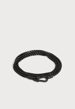 Chaîne métallique tissée noire avec un fermoir mousqueton noir, enroulée sur un fond gris clair.